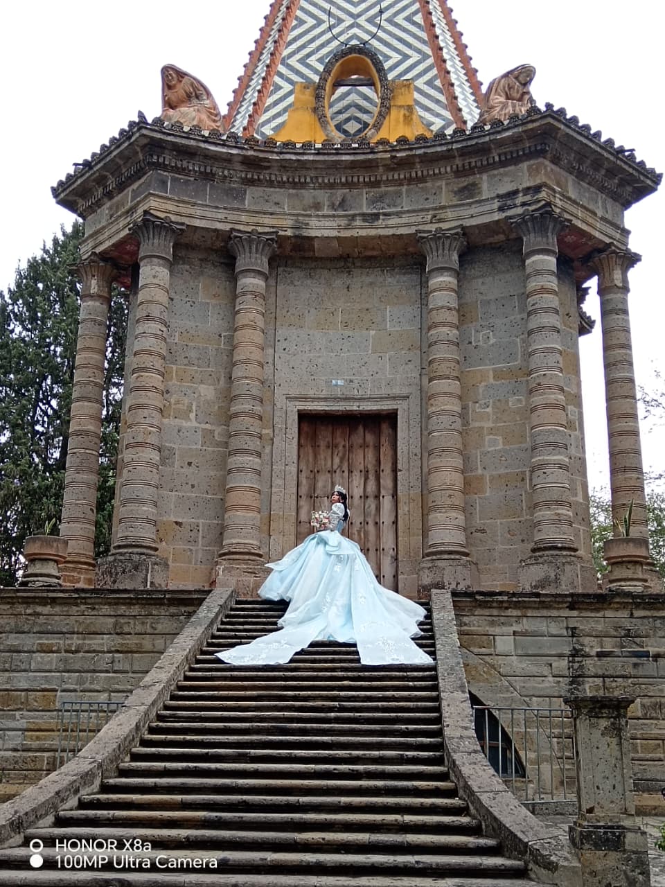 Quinceañera con vestido celeste de princesa en la escalinata de un kiosco antiguo